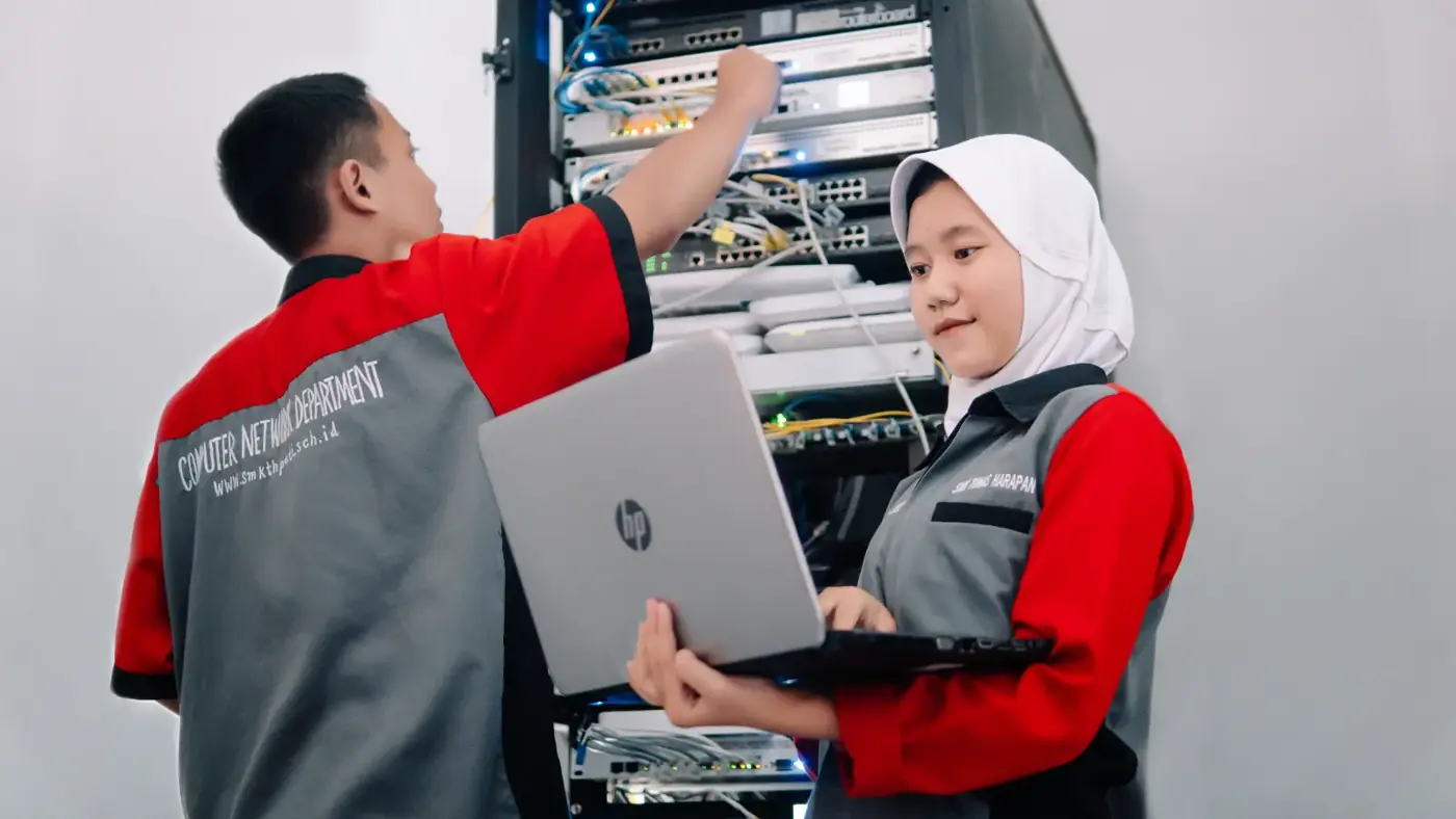 Dua siswa TJKT merapikan perangkat jaringan di rak server sambil memantau lewat laptop